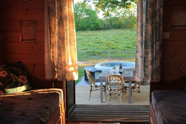 A wooden cabin with a dining area, a hot tub, and a view of a grassy field.