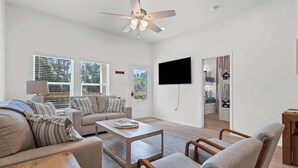A spacious living room with a ceiling fan, a flat-screen TV, and a view of the outdoors through large windows. - Luxury Retreat on Oleander - Unit I (Dauphin Island)