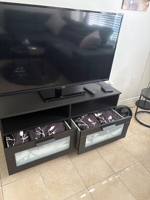 A flat-screen TV on a black entertainment unit with a remote and a smartphone. Below the TV, there are two drawers with patterned fabric inside.