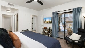A bedroom with a bed, a chair, a desk, a ceiling fan, and a view of the outside.