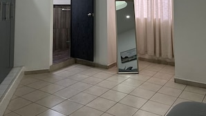 Ein geräumiges Zimmer mit Fliesenboden, einer dunklen Tür, einem großen Spiegel und einem Fenster mit Vorhängen.