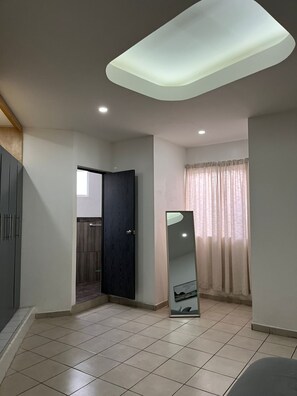 Una habitación amplia con suelo de baldosas, una puerta oscura, un espejo grande y una ventana con cortinas.