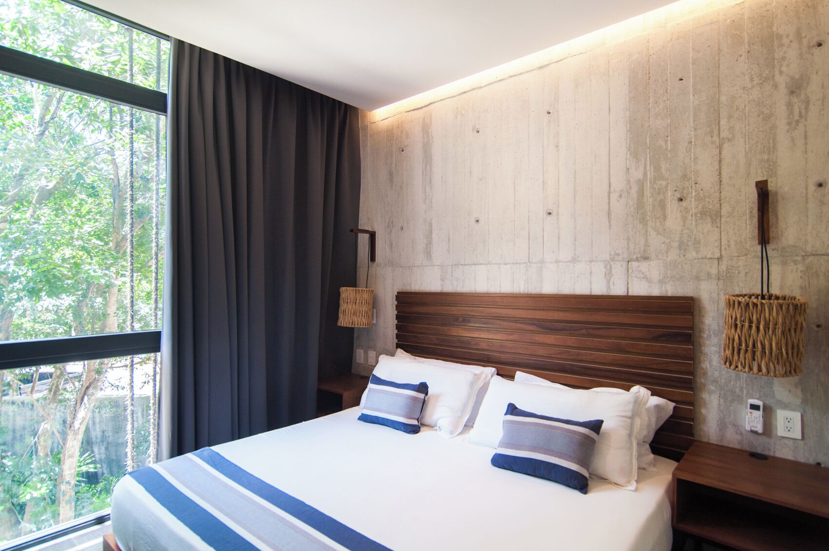 Ein Bett mit Holz-Kopfteil, einer Nachttischlampe und Blick auf Bäume durch ein Fenster.