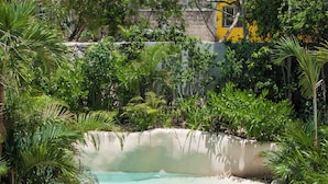 Ein klares Schwimmbecken, umgeben von üppiger Vegetation, mit einem gelben Gebäude im Hintergrund.