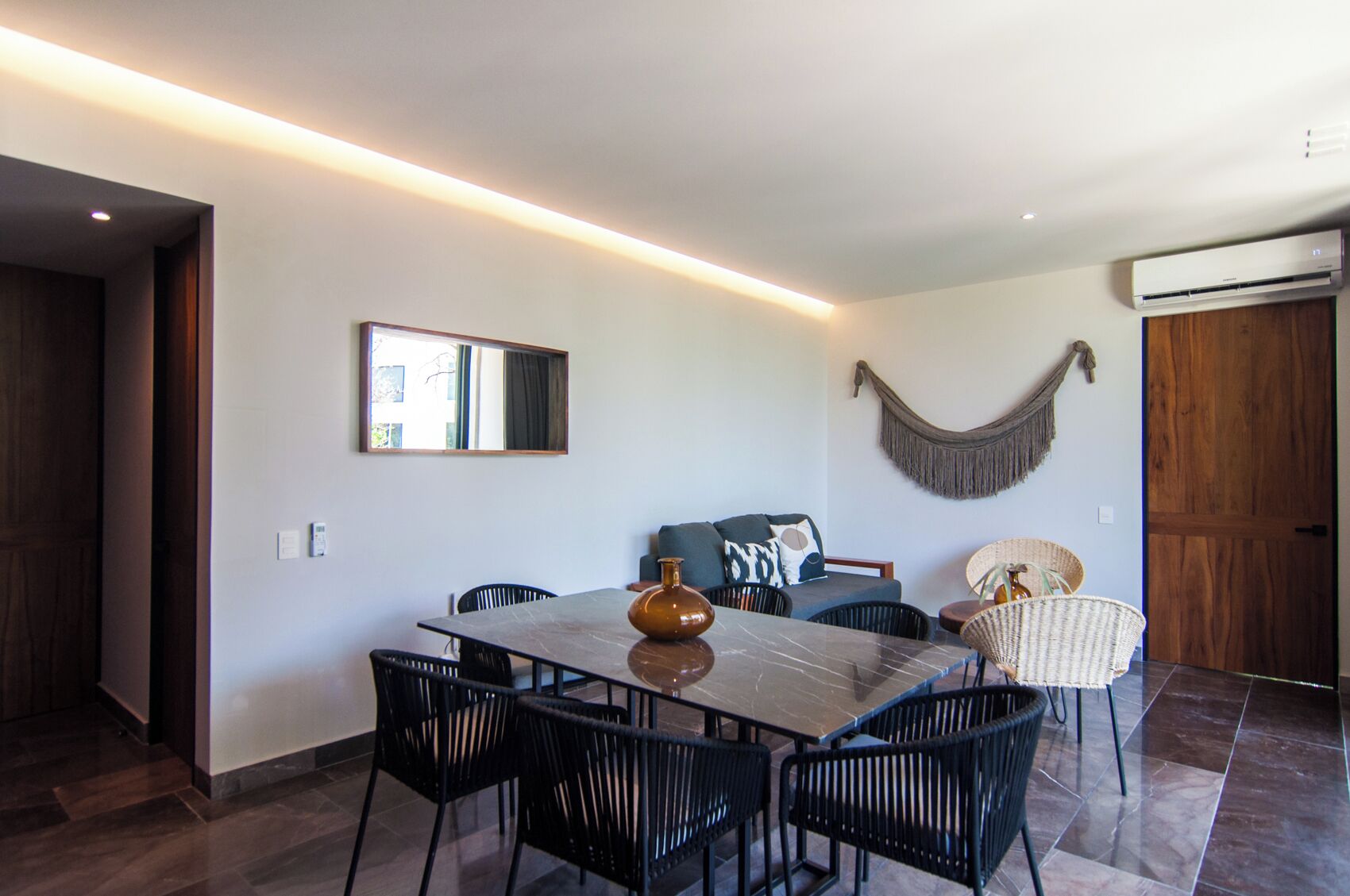 A modern living room with a dining table, black chairs, a sofa, a hanging macrame wall decoration, and a large mirror.
