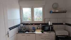 A compact kitchen with white cabinets, a black microwave, a toaster, and a window with a view of greenery. - Studio 4 km from the sea swimming pool and garden Bicycle available for hiking (Villeneuve-lès-Maguelone)