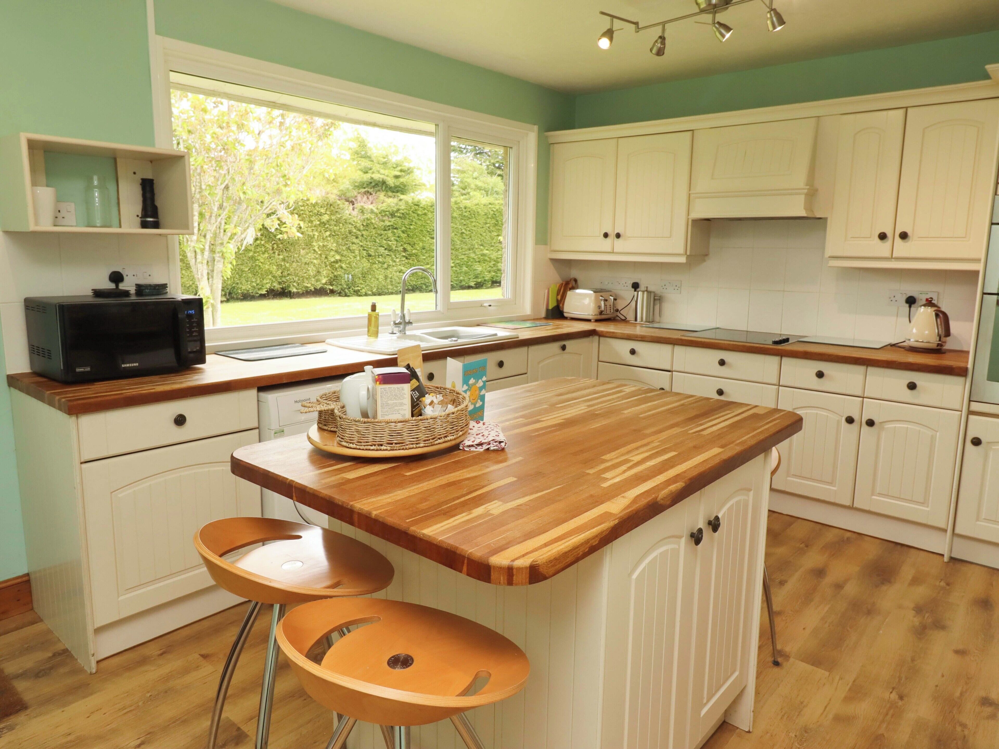 A kitchen with a wooden island, two bar stools, white cabinets, a microwave, and a window with a view of greenery.