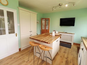 A kitchen with a wooden island, two bar stools, a white refrigerator, a washing machine, a TV on the wall, and a door with a glass panel. - Campden (Alnwick)