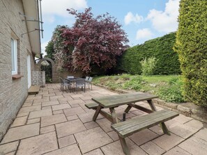 A patio with a table and chairs, a wooden bench, and a garden with a hedge and a tree with red leaves. - Campden (Alnwick)
