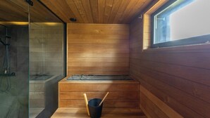 A sauna with wooden walls, a bench, a bucket, and a glass shower enclosure.