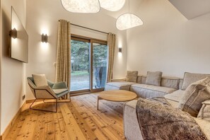 Ein modernes Wohnzimmer mit Holzboden, einer Couch, einem Sessel und einem Couchtisch. Zwei Pendelleuchten und ein großes Fenster mit Vorhängen sind vorhanden.