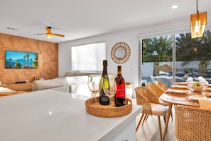 A modern living room with a large island, a sofa, and a dining area with wicker chairs. There are two wine bottles and glasses on the island.
