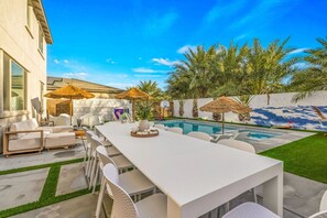 A poolside area with a long white table, white chairs, and a thatched umbrella.