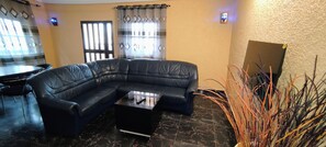 Una sala de estar con un sofá seccional de cuero negro, una mesa de centro de vidrio, una mesa de comedor con sillas, una ventana con cortinas y una lámpara de pared.