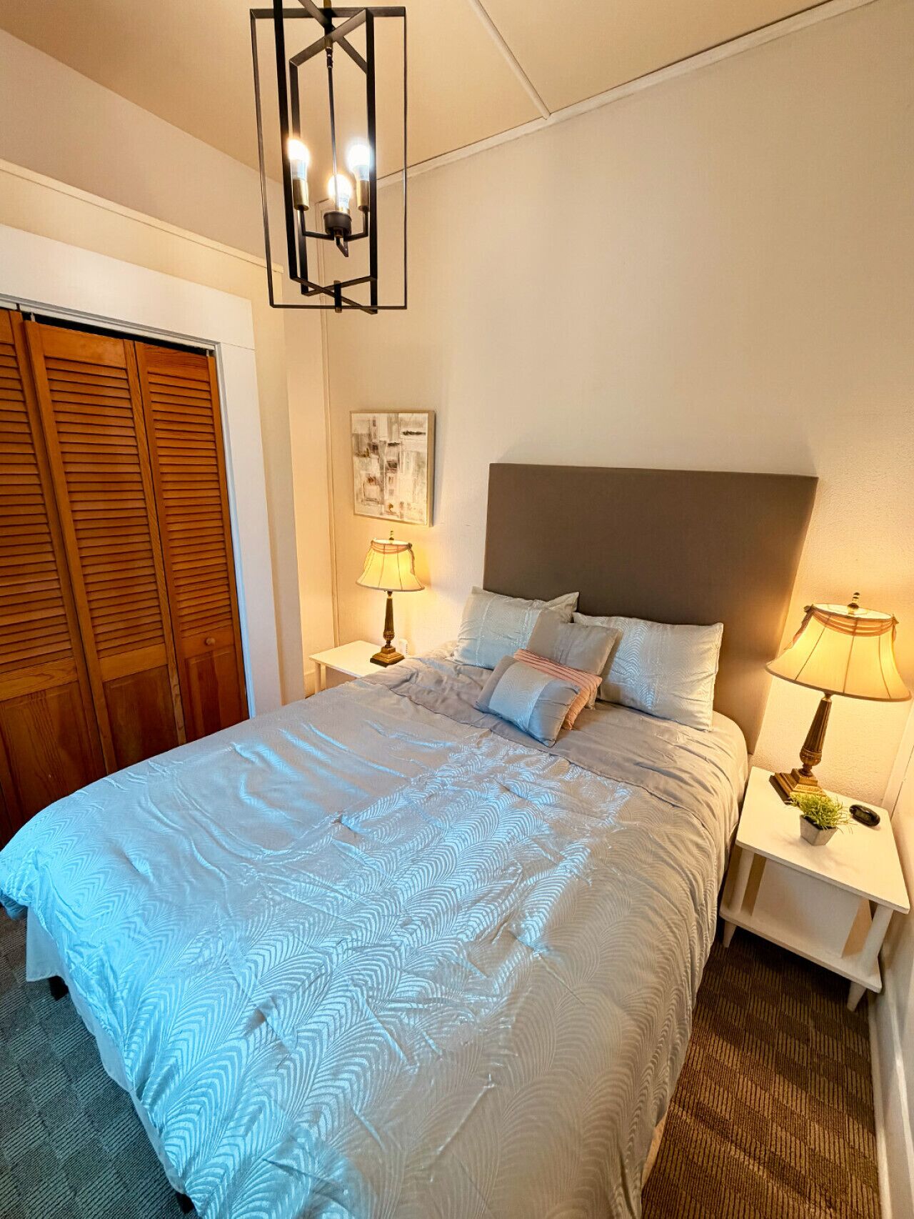 A neatly made bed with a patterned duvet, two bedside lamps, a wooden wardrobe, and a wall-mounted light fixture.
