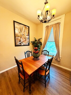 A dining room with a wooden table, four chairs, a chandelier, a painting, a potted plant, and a window with curtains.