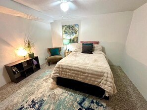 Ein Schlafzimmer mit Bett, Sessel, Nachttisch, Lampe, Bücherregal und einem Bild an der Wand.