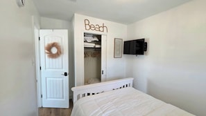 Ein Schlafzimmer mit weißer Tür, einem weißen Bett, einem Kleiderschrank mit Kleidung und einem Fernseher an der Wand. Über dem Kleiderschrank hängt ein Schild mit der Aufschrift 'Beach'.