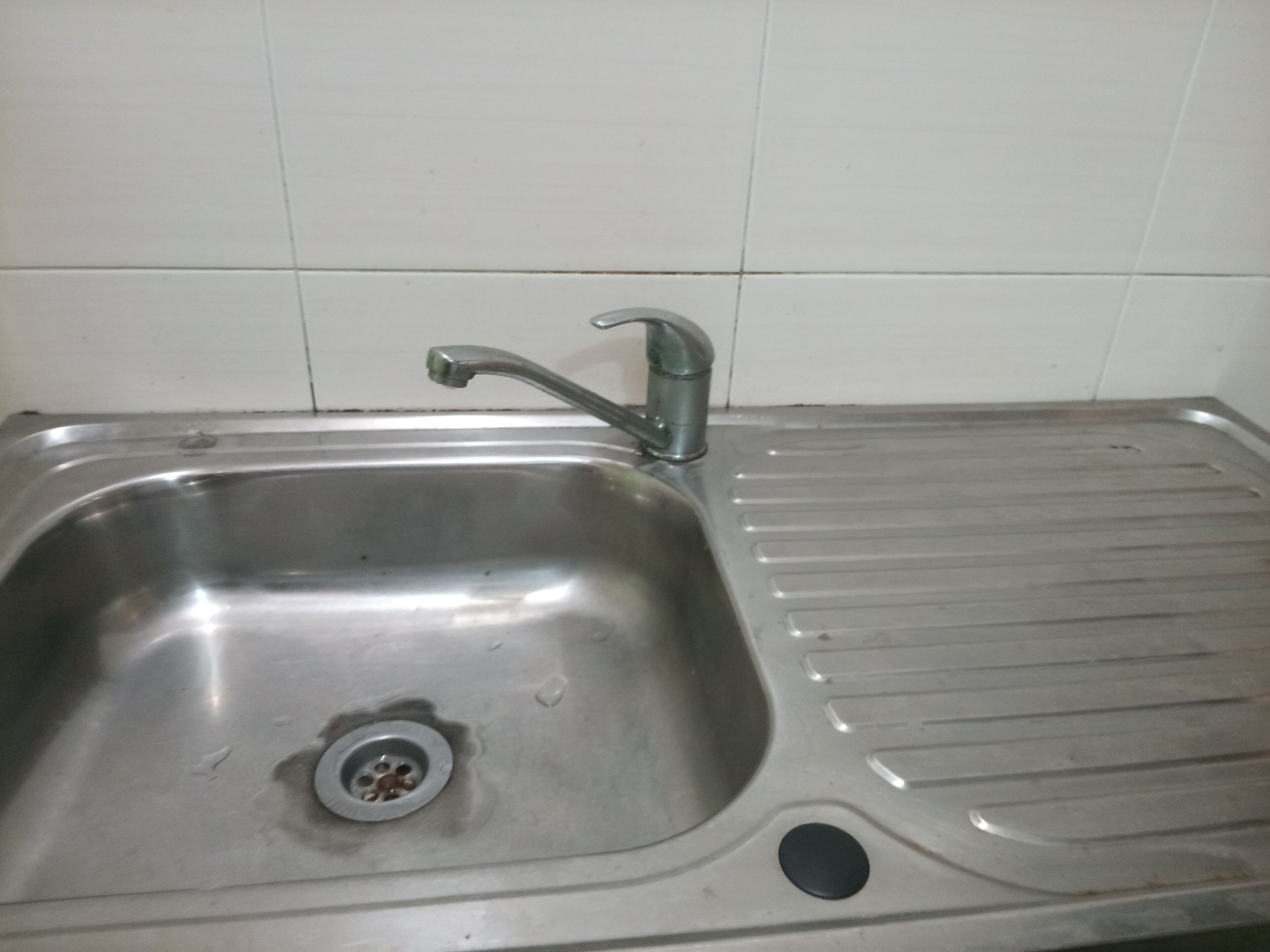 Un lavamanos de acero inoxidable con un solo grifo, un desagüe y un objeto redondo y negro a la derecha.