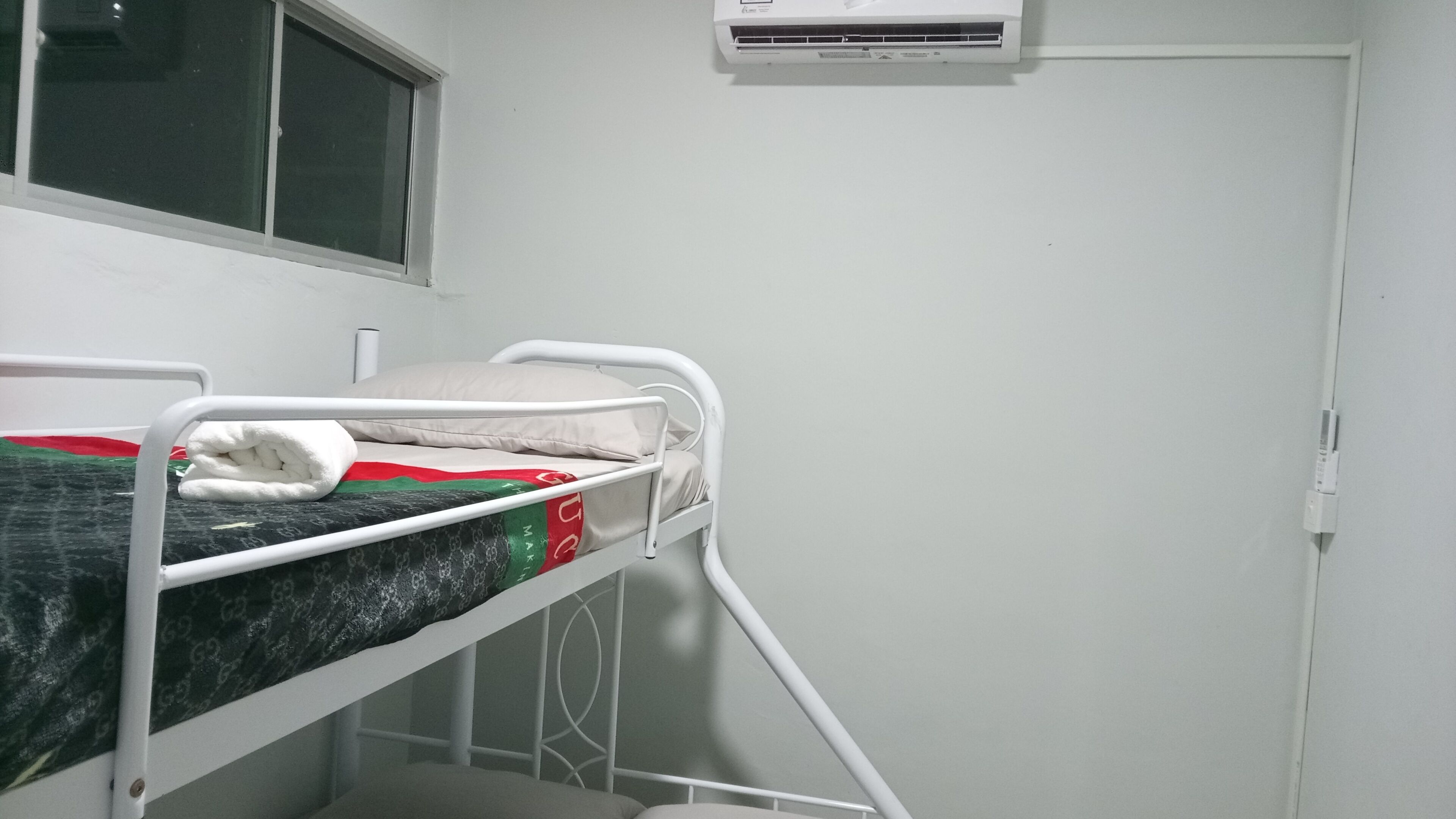 Un letto singolo con struttura in metallo bianco, una finestra con persiane e un condizionatore d'aria a parete.