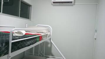 Un letto singolo con struttura in metallo bianco, una finestra con persiane e un condizionatore d'aria a parete.
