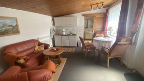 A cozy living room with a leather sofa, a small round table, and a dining area with wooden chairs. A kitchen with white cabinets and a wooden ceiling is visible in the background. - Vacation apartment with green garden view (Ahrenshoop)