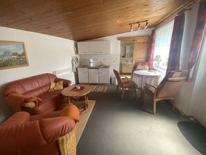 Ein gemütliches Wohnzimmer mit einer Ledersofa, einem kleinen runden Tisch und einem Essbereich mit Holzsesseln. Im Hintergrund ist eine Küche mit weißen Schränken und einer Holzbalkendecke erkennbar.