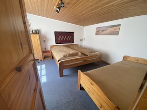 A room with a wooden ceiling, a bed with a beige blanket, a small table with a lamp, and a wall-mounted picture.