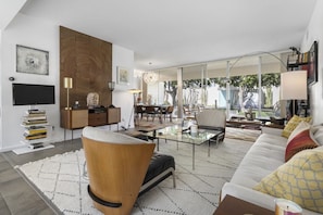 A modern living room with a glass coffee table, a sofa, a wooden accent chair, and a dining area with a chandelier. - Large, airy mid-century condo at 7 Lakes golf club (Palm Springs)