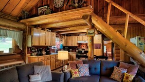Interior rústic d'una cabana amb escala de fusta, zona de dinar, cuina i saló amb sofà i coixins.