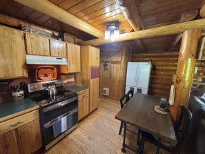 A wooden cabin interior with a kitchen area featuring a stove, oven, refrigerator, wooden cabinets, and a dining table with chairs.