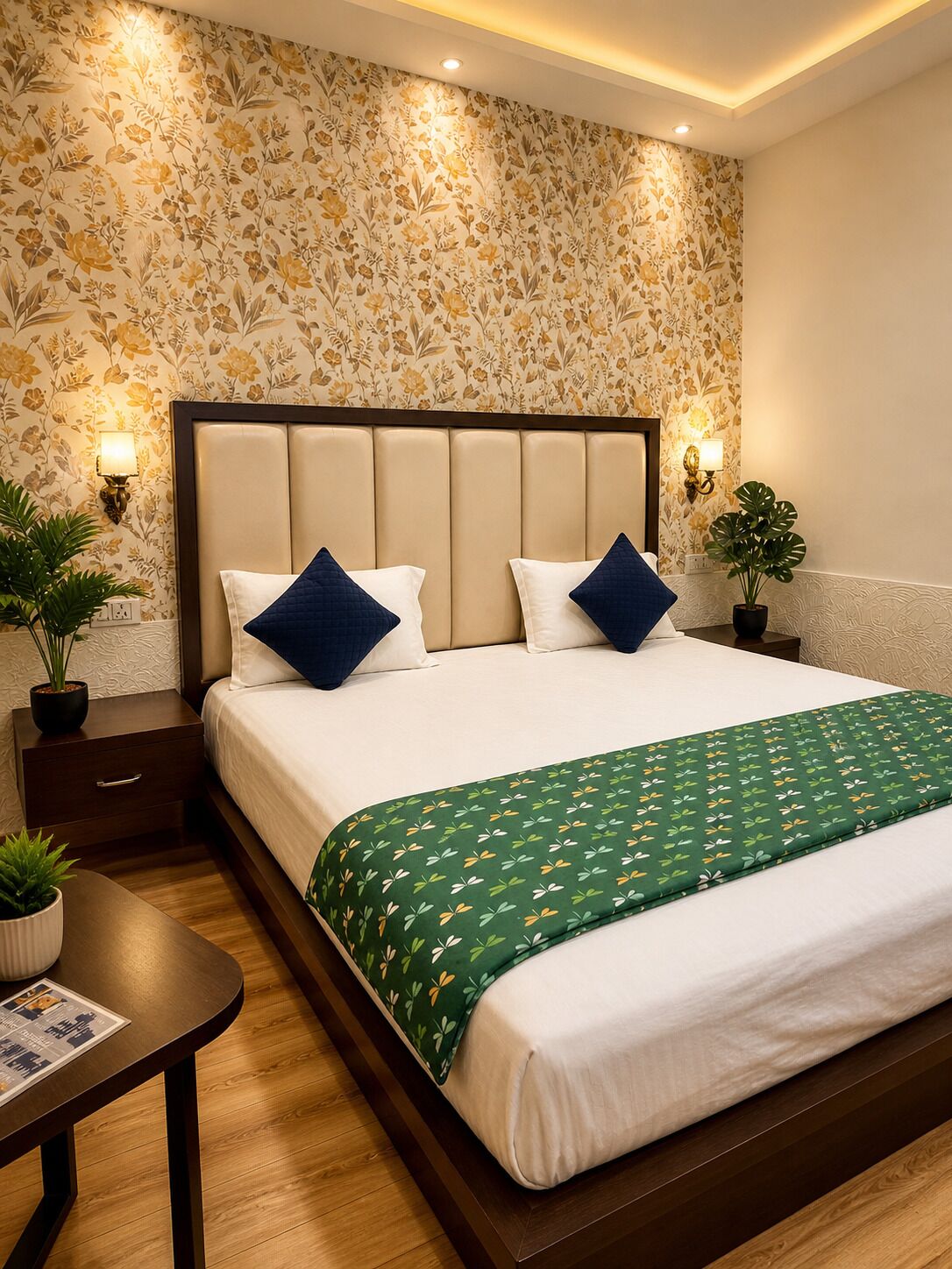 Un dormitorio ordenado con papel tapiz floral, cabecera capitonada, dos mesitas de noche con lámparas, colcha estampada verde y una pequeña planta en maceta sobre la mesita.