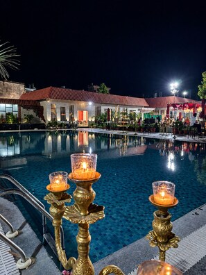 A nighttime pool area with a clear view of the pool, a few lit candles on ornate stands, and a building with red-tiled roofs in the background. - The Sparkle  (Jaipur)