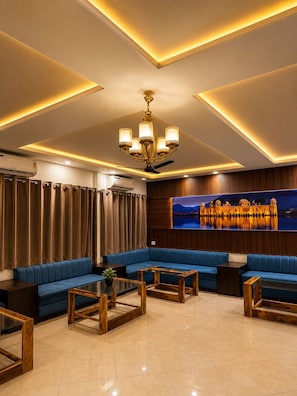 A modern hotel lobby with a blue sofa set, wooden coffee tables, a chandelier, and a large wall-mounted TV displaying a scenic view. - The Sparkle  (Jaipur)