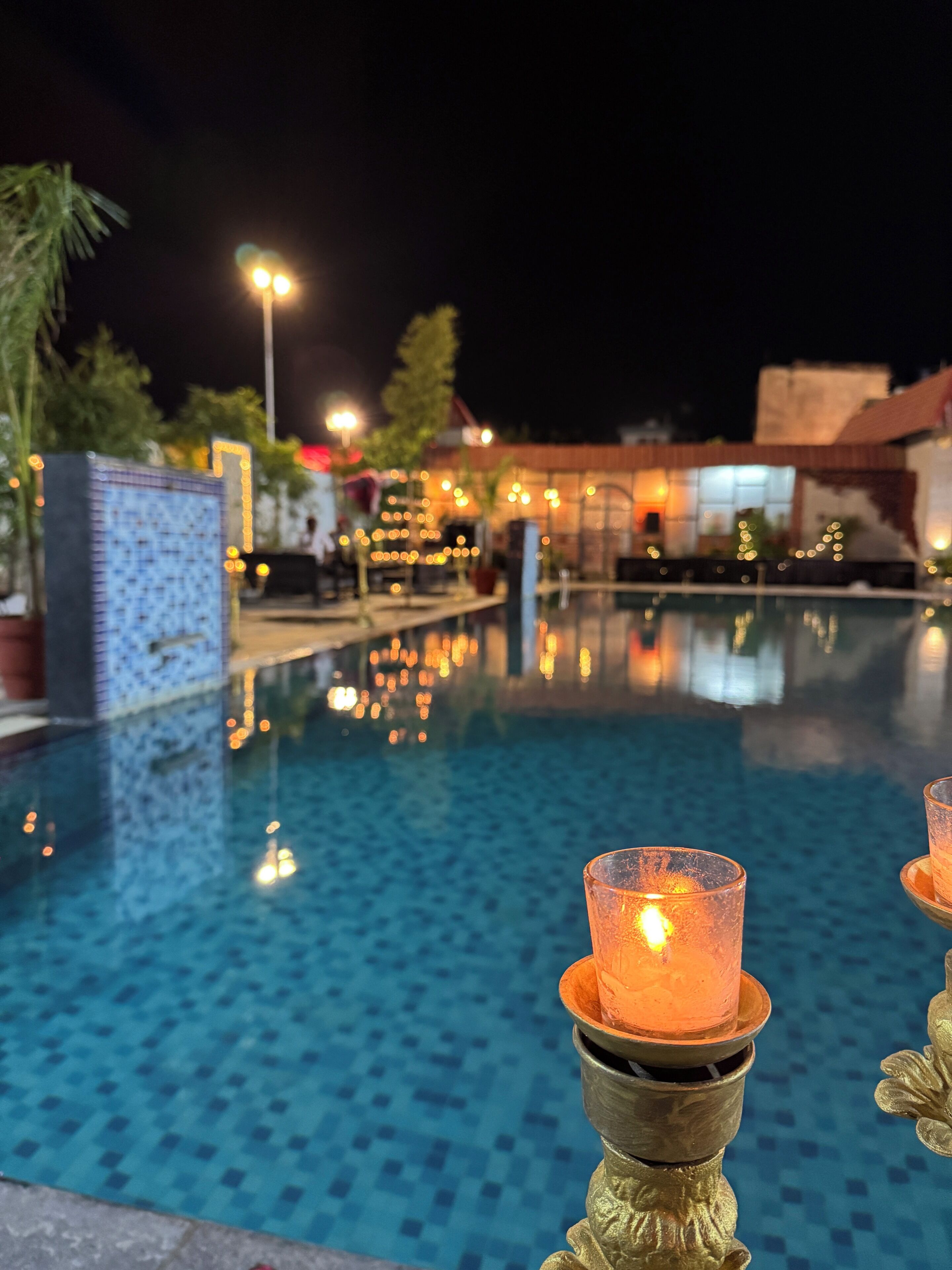 Zona de piscina nocturna con vista despejada del agua, algunas velas encendidas y un edificio distante.
