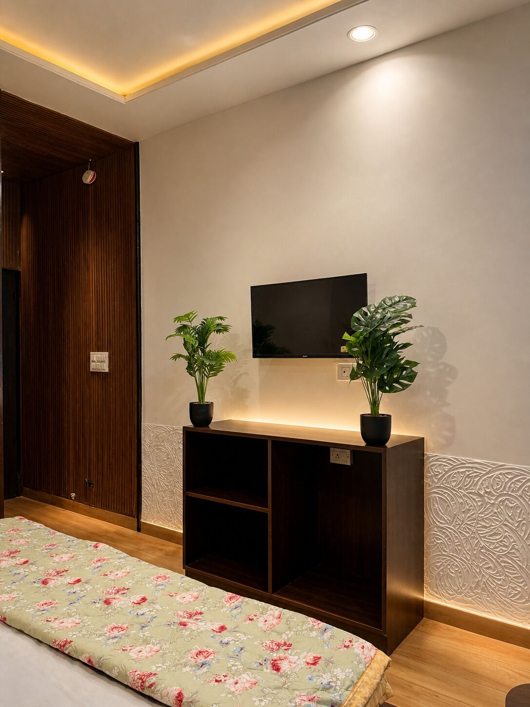 Quarto com uma cama, televisão na parede e duas plantas em vasos.