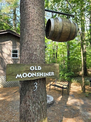 En träskylt med texten ”OLD MOONSHINER” och siffran ”3” fäst på ett träd, en metalltunna som hänger från en gren och en bänk i ett skogsområde.