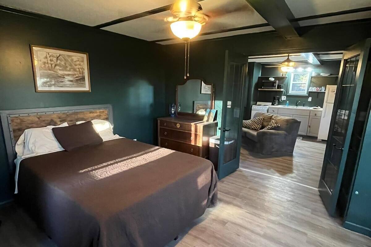 Ein Schlafzimmer mit Bett, Kommode, Nachttisch, Deckenventilator und Blick in einen Küchenbereich.