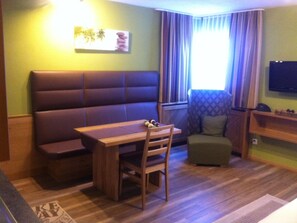 Ein Zimmer mit einer Couch, einem Holztisch, einem Sessel, einem kleinen Hocker, einem Fernseher und einem Fenster mit Vorhängen.