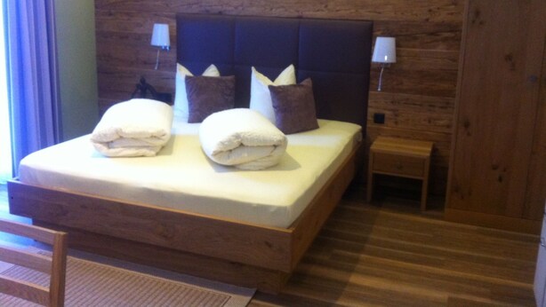 A wooden-framed bed with pillows and a bedside table in a room with wooden walls and a window with curtains. - Apart Bellezza, (Samnaun-Dorf) by Interhome (Samnaun)
