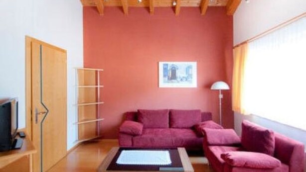 Una stanza con soffitto in legno, parete rossa, divano, tavolino da caffè, televisore e finestra con tende.