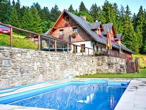 A wooden chalet with a stone retaining wall, a swimming pool, and a forested backdrop. - Penzion Exnar by Interhome (Strážné)