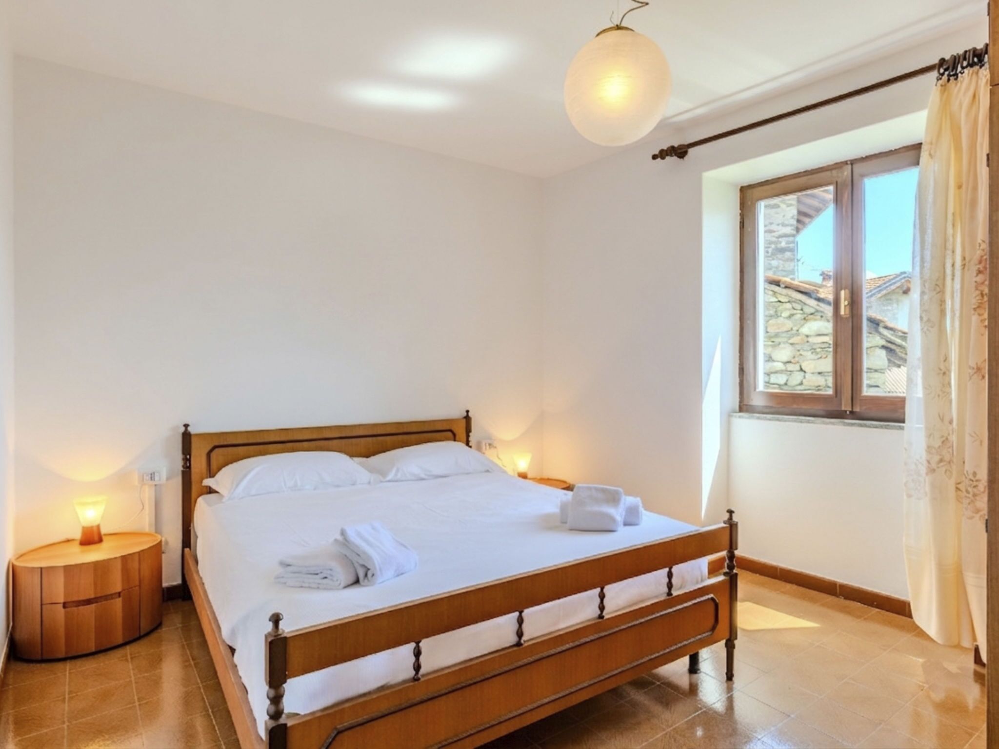 A bedroom with a wooden bed, bedside lamps, a window with curtains, and a view of buildings outside.