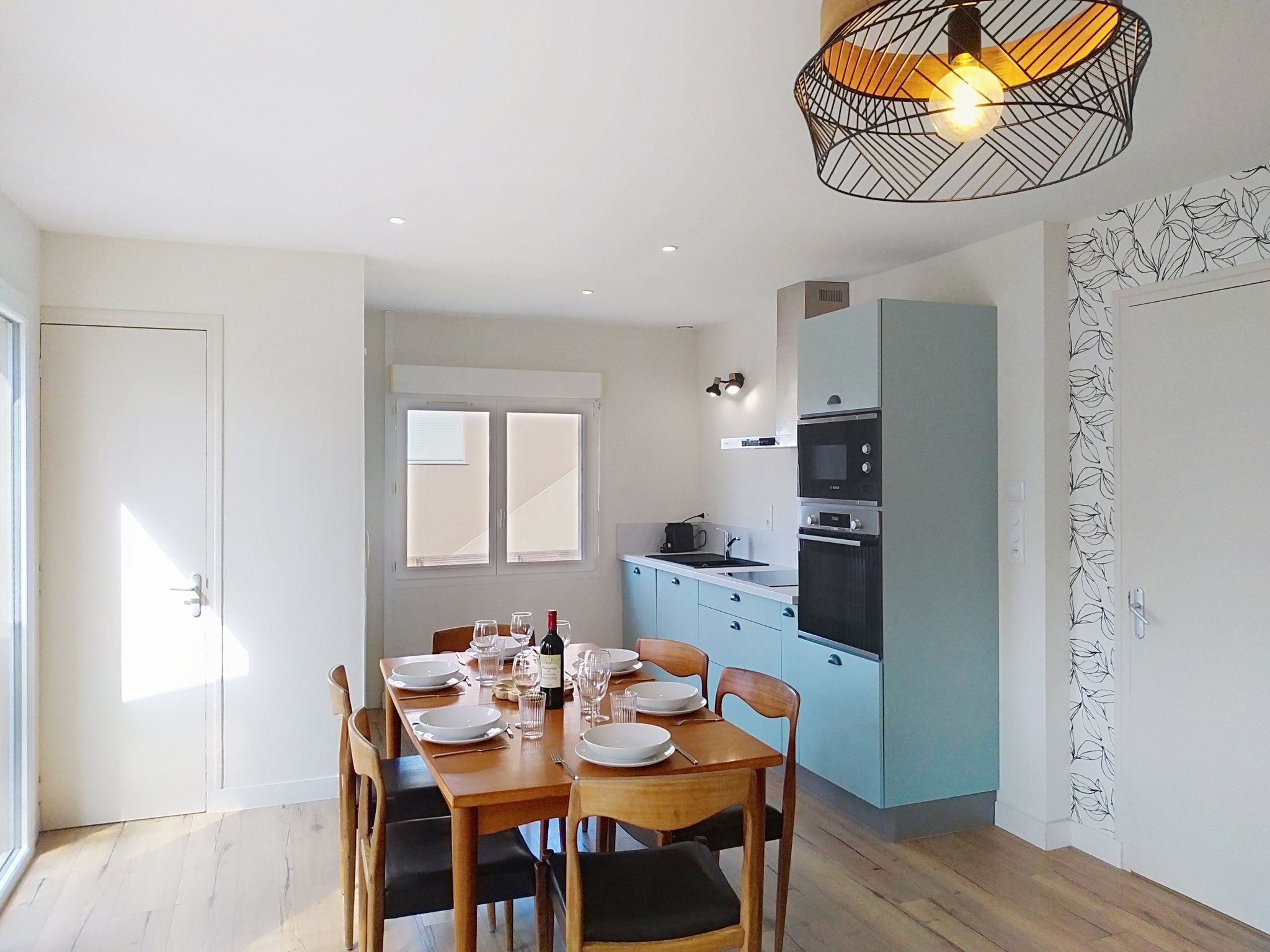 Un comedor con una mesa dispuesta para cuatro personas, una cocina con armarios azules y una lámpara en el techo.