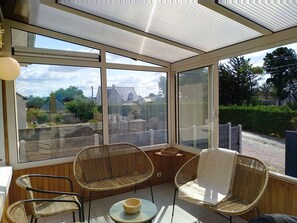 A sunroom with large windows, wicker chairs, a small table, and a view of a residential area. - Dolce Vita by Interhome (Agon-Coutainville)
