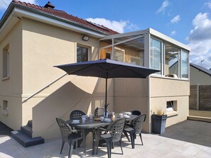 Une terrasse avec une table et des chaises, un grand parasol, et une maison avec un toit en tuiles et une véranda en verre.