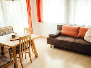 En stue med et træbord, to stole, en sofa med orange puder og et vindue med lette gardiner.