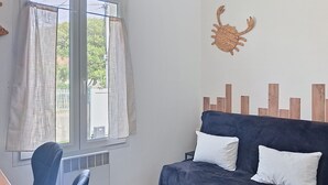 Um quarto com uma janela, uma cadeira azul, um sofá azul escuro com dois travesseiros brancos e uma decoração de parede com um caranguejo tecido.