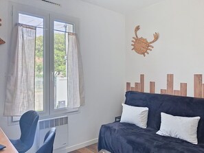 Une pièce avec une fenêtre, une chaise bleue, un canapé bleu foncé agrémenté de deux coussins blancs, et une décoration murale représentant un crabe tissé.