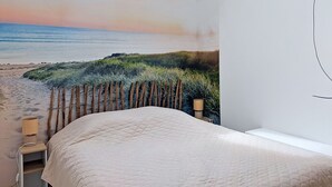 Quarto com mural de praia, uma cama, mesa de cabeceira e uma janela.